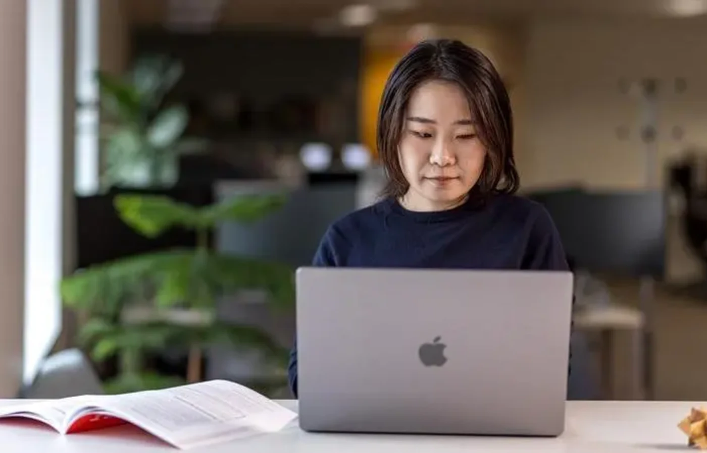 Asian woman sitting at a Macbook with an open book next to her.