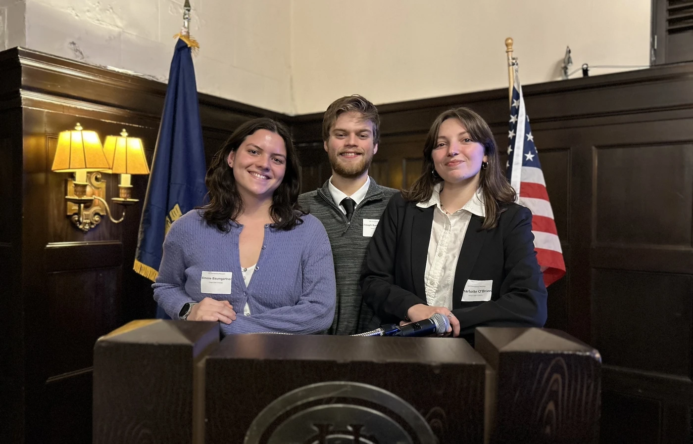 Three students smile at a podium flanked by the U.S. and Oregon state flags.