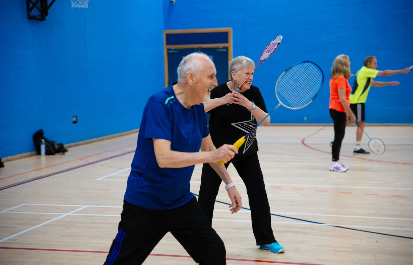 Two elderly white people holding badminton rackets and smiling in a gym.
