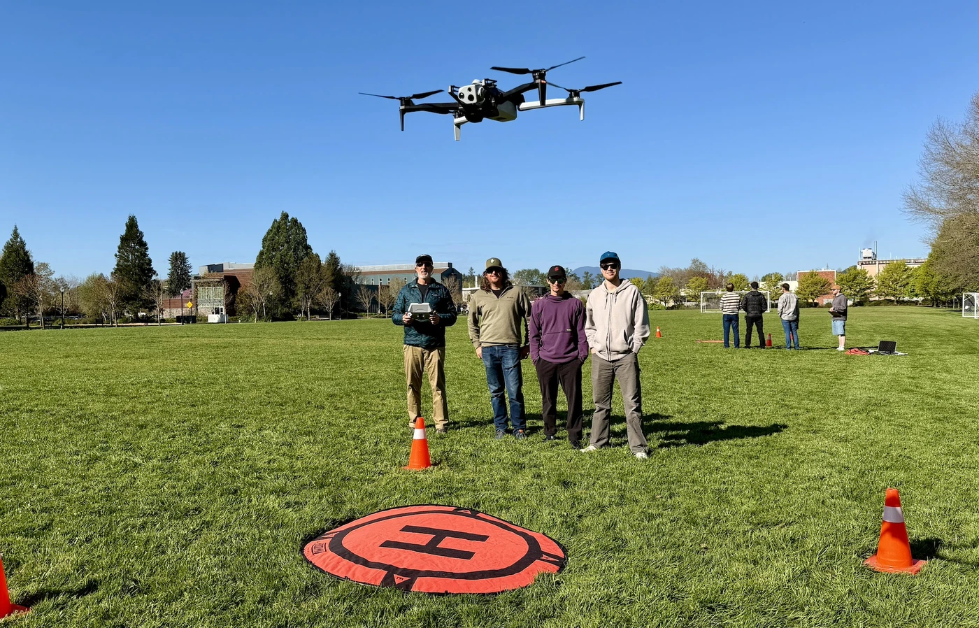Four men stand side by side on Peavy Field while a drone hovers in front of them over an orange landing pad.