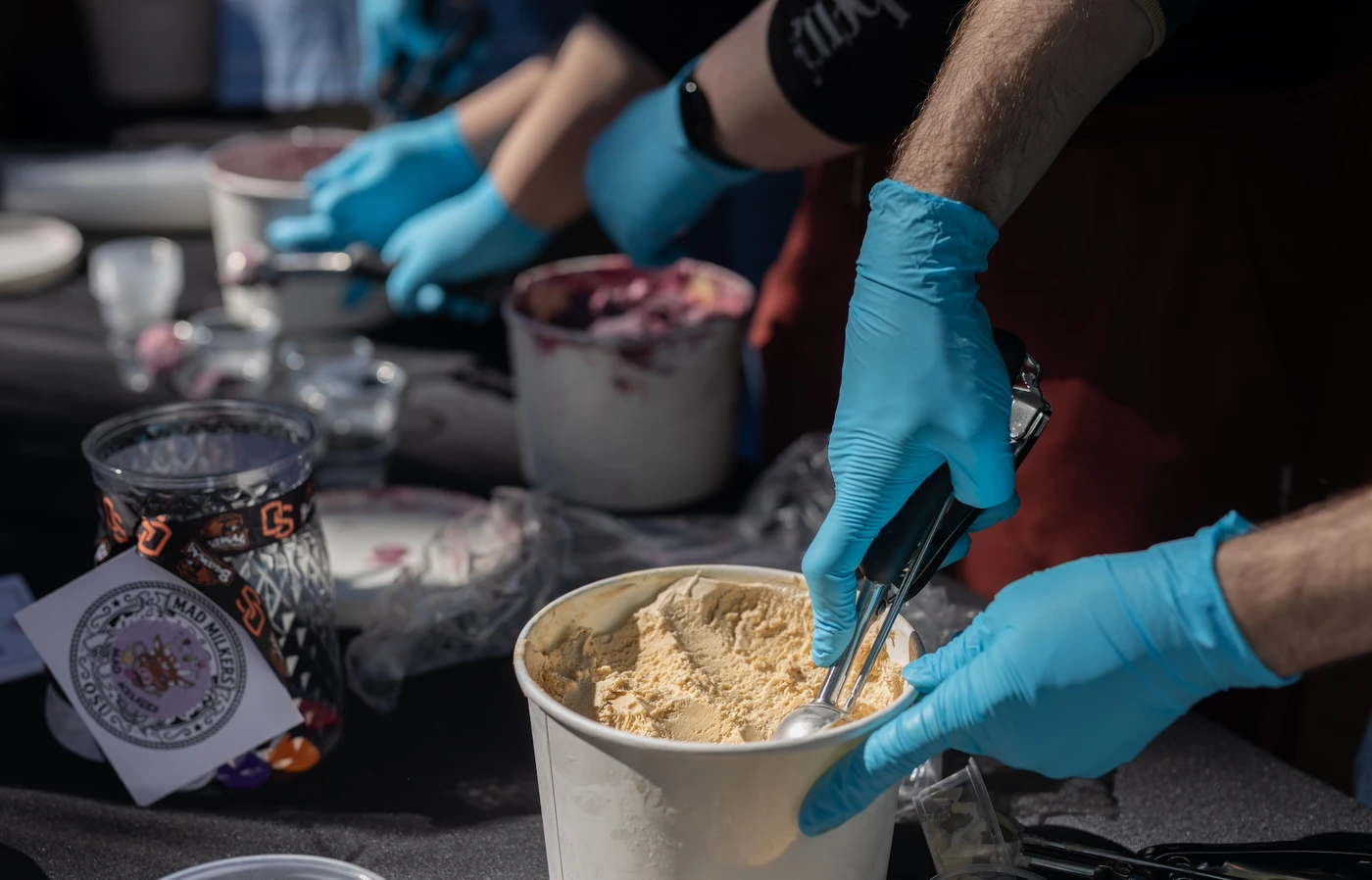 Blue-gloved hands scoop ice cream out of a white carton.