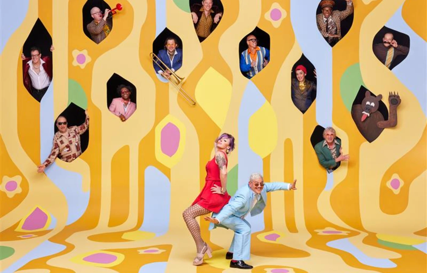 A creative photo of the band Pink Martini, with the lead pianist and singer standing on a psychedelic yellow painting in the foreground and various band members popping out of windows above them.