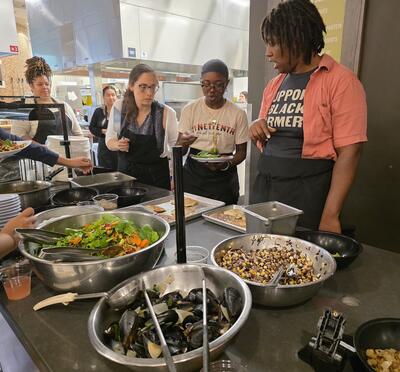 A Black woman teaches a cooking class with several large bowls of food on a dining hall kitchen counter.