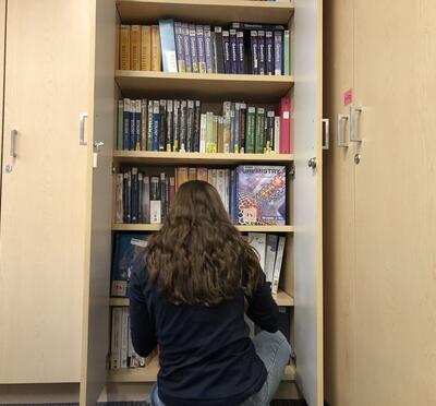 A young woman with long dark hair crouches in front of bookshelves, back to the camera.