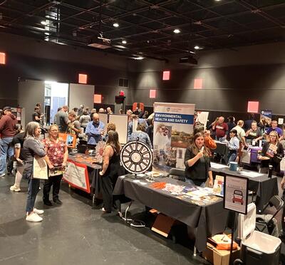 A large collection of people and various decorated tables at University Day in the black box theater of PRAx.