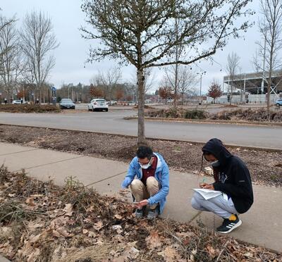 Two students in medical masks crouch on the sidewalk next to the dirt where they have installed a seismometer.