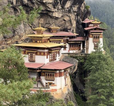 A white, red and gold temple built into the side of a cliff in the tree-covered mountains of Bhutan.