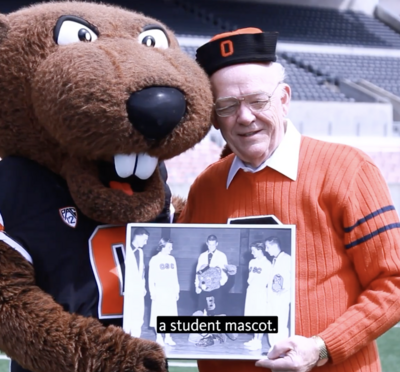 The Benny Beaver mascot standing next to an elderly man in an orange sweater, who was the first person to embody Benny.