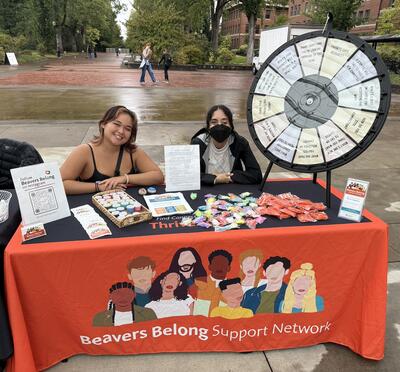 Two students sitting at a Beavers Belong Support Network table during an outdoor tabling event.