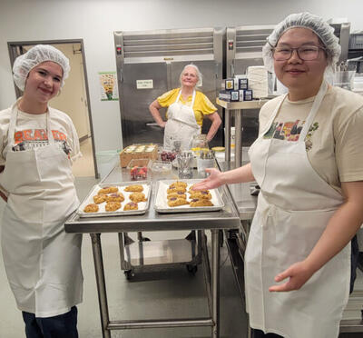 Three students in hairnets and white aprons pose with trays of scones in the Beaver Classic kitchen.