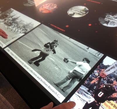 A story displayed on the OSU 150 touch table shows an old black-and-white photo of Benny the Beaver on a baseball field.