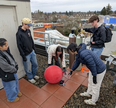 A cluster of five people watch and lean down as the red weather balloon is inflated on the roof.