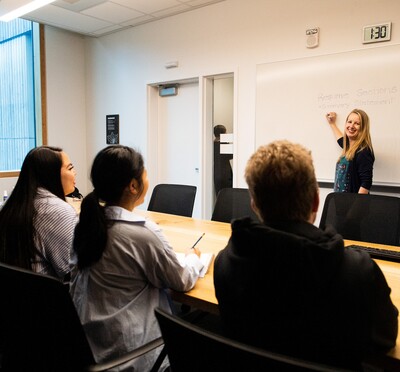 A blonde woman stands at the whiteboard explaining something to a group of three people sitting across from her.