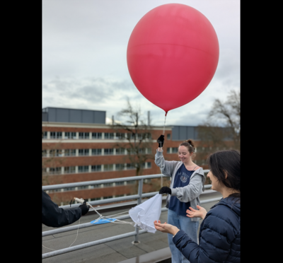 A student holds onto the tail of a big red weather balloon on the roof of an OSU building pre-launch.
