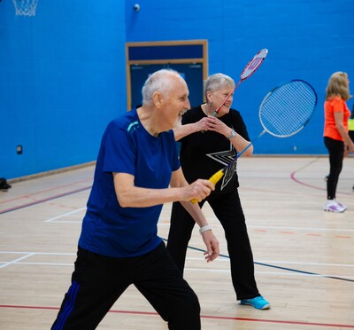 Two elderly white people holding badminton rackets and smiling in a gym.