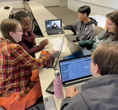 A group of business students sit around a narrow table with their laptops working together.