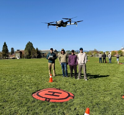Four men stand side by side on Peavy Field while a drone hovers in front of them over an orange landing pad.