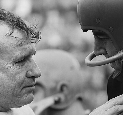 Black-and-white photo of a grizzled athletic trainer addressing a football player.