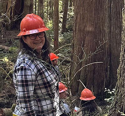 Woman in orange hard hat and flannel shirt smiling in the forest.