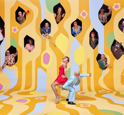 A creative photo of the band Pink Martini, with the lead pianist and singer standing on a psychedelic yellow painting in the foreground and various band members popping out of windows above them.