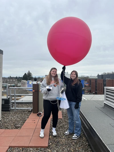 Two female students smile as they hold the big red weather balloon over their heads.