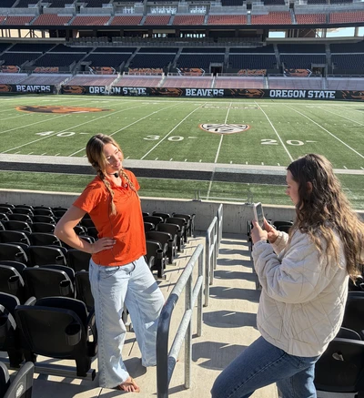 Blonde-haired Emily Engelke-Ryan in an orange shirt posing in the stands of Reser Stadium while another girl takes her photo.