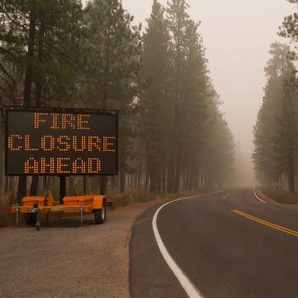 Image shows a large electronic road sign to the left of a highway announcing "Fire closure ahead." The road winds through a smoke-filled forest.