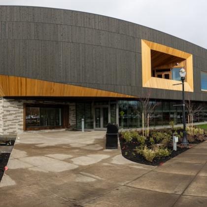 Wide-angle shot of the main entrance of PRAx, a new building on OSU's campus with a dark gray overhanging second story, large inset windows trimmed in blond wood and a wall of windows showing the first-floor lobby. 