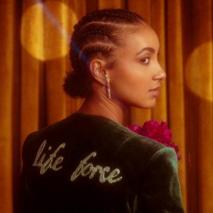 Image shows jazz artist esperanza spalding in front of a copper-colored curtain; she's wearing a green velvet jacket with "life force" written across the back, and she's looking at the camera over her right shoulder. 