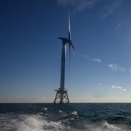 Image shows a series of 5 massive offshore wind turbines in open ocean under a glaring sun; the first turbine is close by in the foreground and the others look progressively smaller as they are farther away.
