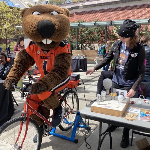 Benny Beaver riding the Sustainability Office's Bike Generator, using it to power a lightboard on a nearby table.