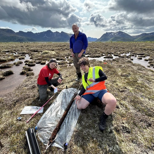 Two people crouching and one person standing during fieldwork in front of a mountain range in Sweden.