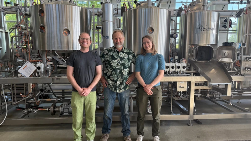 A young man, an older man, and a young woman smile as they stand in front of the large metal vats in OSU's pilot brewery.