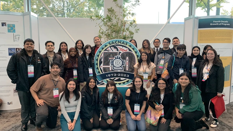 A group of mostly Hispanic and Indigenous students poses around the SACNAS 1973-2023 display sign at a conference.