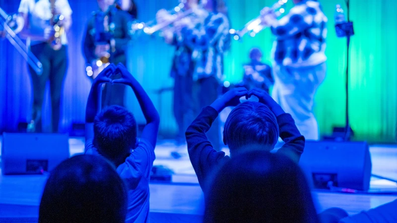 Two small children in the foreground hold up heart-hands to the musicians performing onstage, awash in blue light.