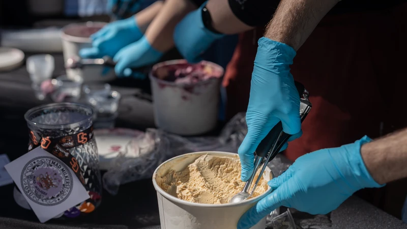 Blue-gloved hands scoop ice cream out of a white carton.