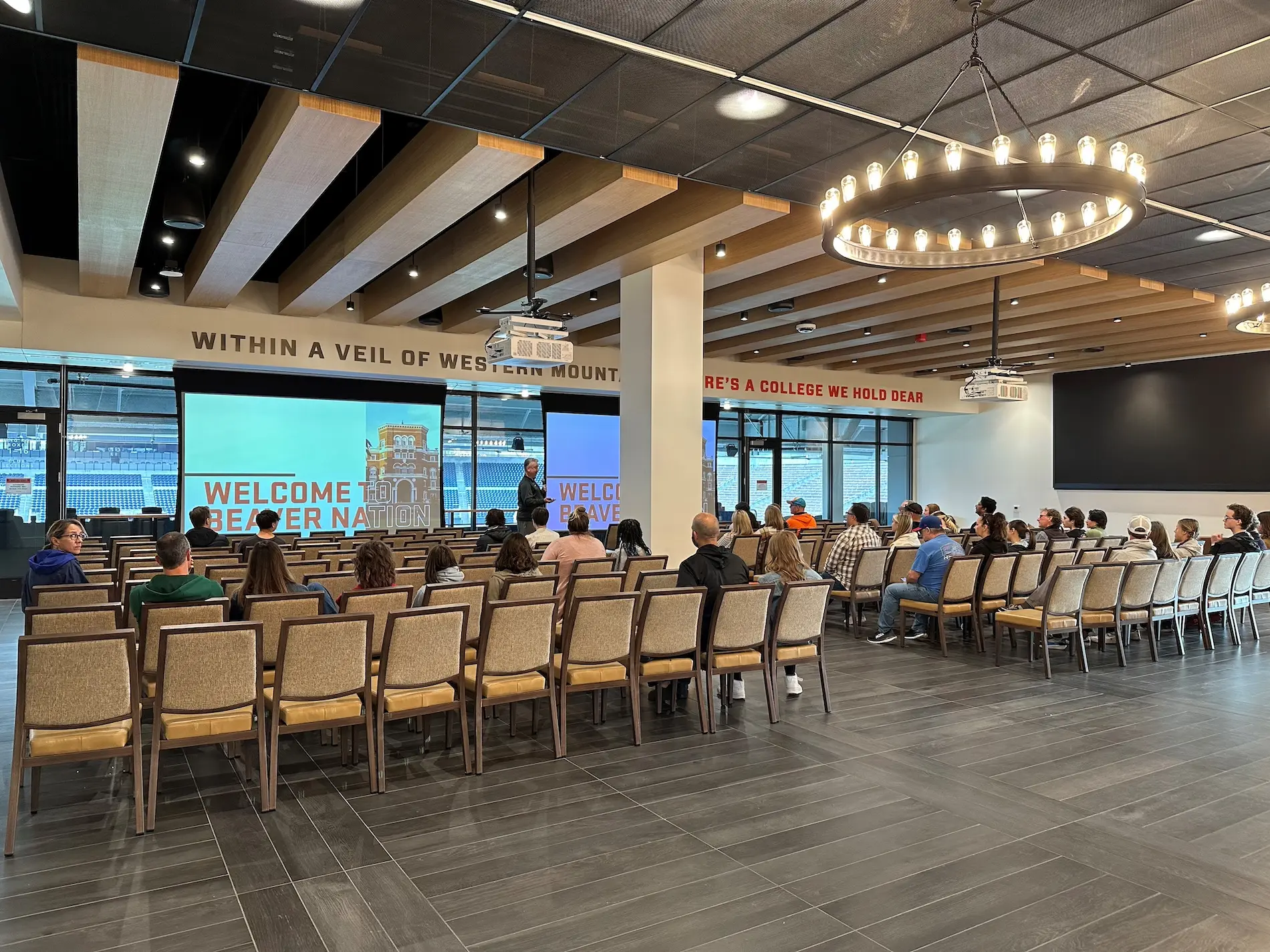 Seen from behind, a group of people sit in chairs in the Welcome Center's presentation area, watching the projector screen.