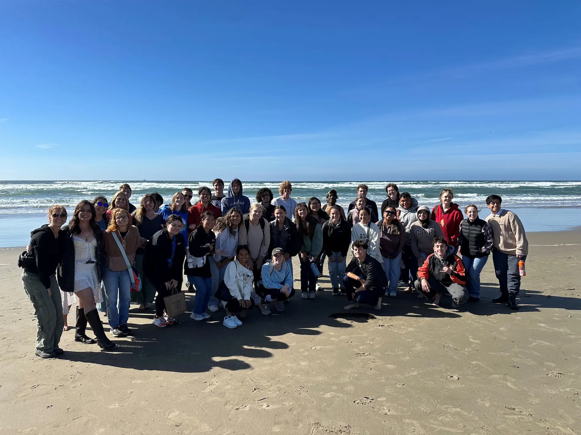 A large group of students smile for the camera at the beach in Newport on a sunny day.