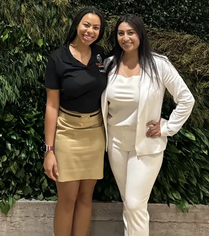 Two women smiling in front of a leafy wall. 
