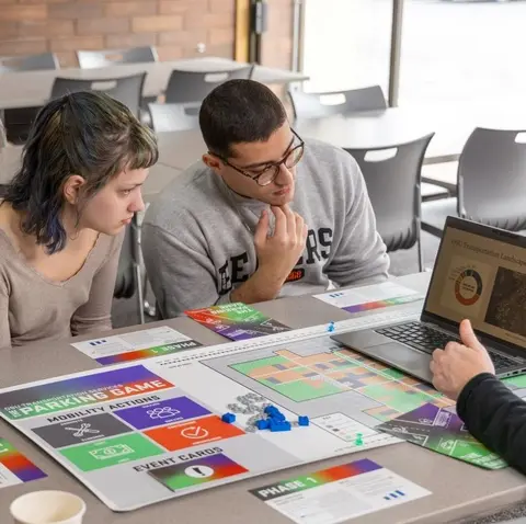 Two students lean over a table playing the Parking Game with cards and game board in front of them.