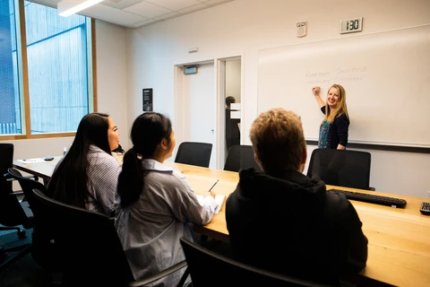 A blonde woman stands at the whiteboard explaining something to a group of three people sitting across from her.