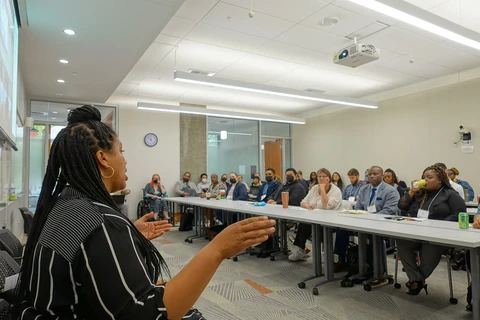 A Black woman in the foreground addresses a classroom of attendees.
