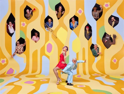 A creative photo of the band Pink Martini, with the lead pianist and singer standing on a psychedelic yellow painting in the foreground and various band members popping out of windows above them.