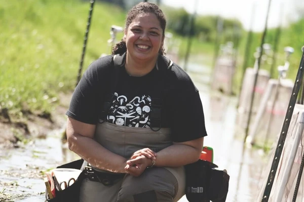 Image shows Rahiza De Thomas in fishing waders knee-deep in a murky brown agricultural channel.