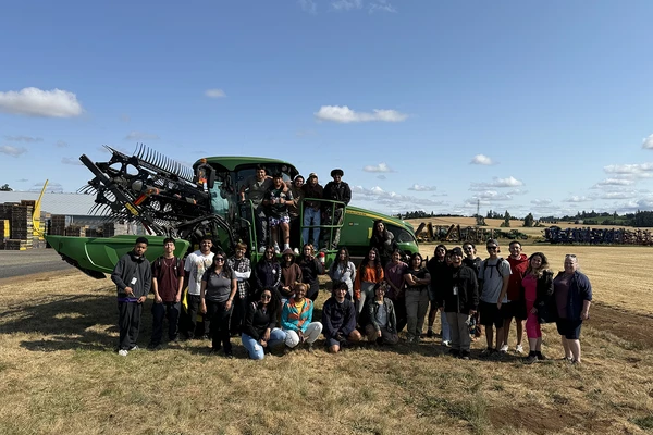 Group of students stand in front of a large piece of farm equipment in a field.