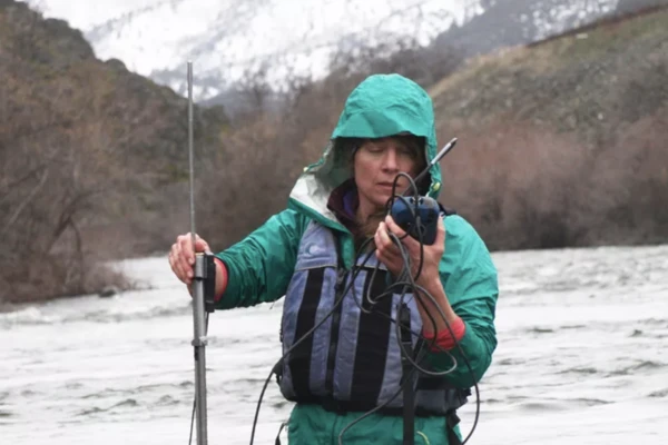 Researcher Desiree Tullo stands in the middle of a river, holding monitoring equipment and wearing a raincoat and life jacket.