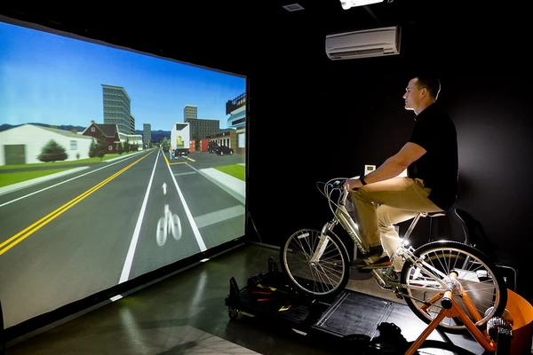 A test subject rides a stationary bike in front of a large screen showing a projection of a bike lane on a city street.