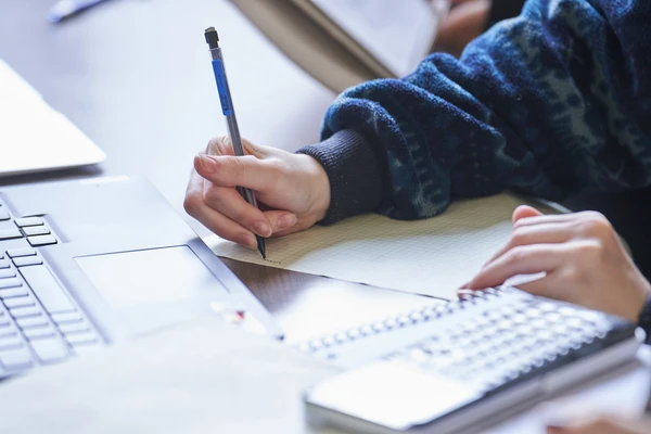 Stock photo of hands writing on paper near a laptop.