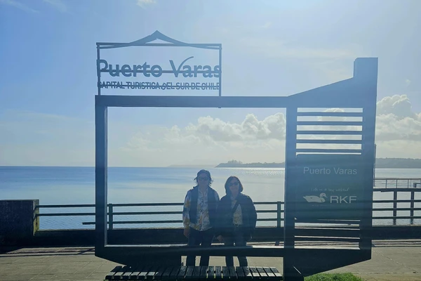 Two women stand under a Puerto Varas sign with the ocean in the background