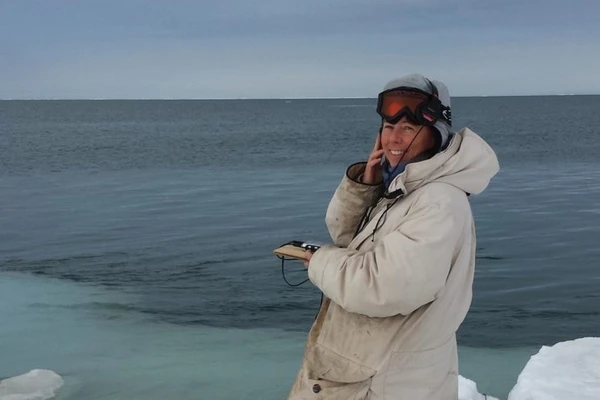 Researcher Kate Stafford stands on edge of the ice in Alaska, wearing a long parka and holding a hydrophone used to record underwater sound.